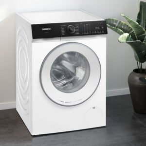 Le lave-linge à chargement frontal SIEMENS WG56H2A5FR se trouve dans une buanderie avec une plante, un panier et des vêtements soigneusement pliés à proximité.