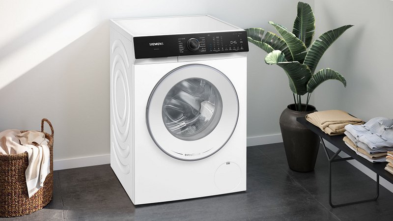 Le lave-linge à chargement frontal SIEMENS WG56H2A5FR se trouve dans une buanderie avec une plante, un panier et des vêtements soigneusement pliés à proximité.