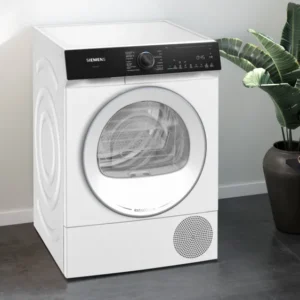 Le sèche-linge SIEMENS WQ46H2CAFR se trouve dans une buanderie moderne avec une plante, un panier à linge et des vêtements soigneusement pliés sur le banc.