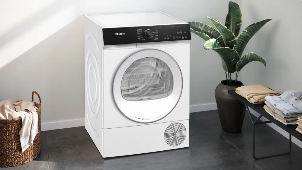 Le sèche-linge SIEMENS WQ46H2CAFR se trouve dans une buanderie moderne avec une plante, un panier à linge et des vêtements soigneusement pliés sur le banc.