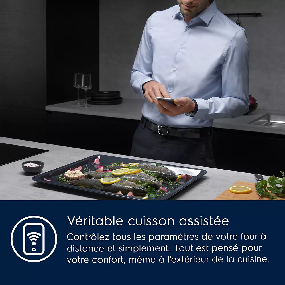 Un homme utilise son téléphone près d'un plateau de poisson et de légumes dans une cuisine moderne, vérifiant que le four ELECTROLUX COD6P46X0 (Copie) donne des résultats de cuisson parfaits.