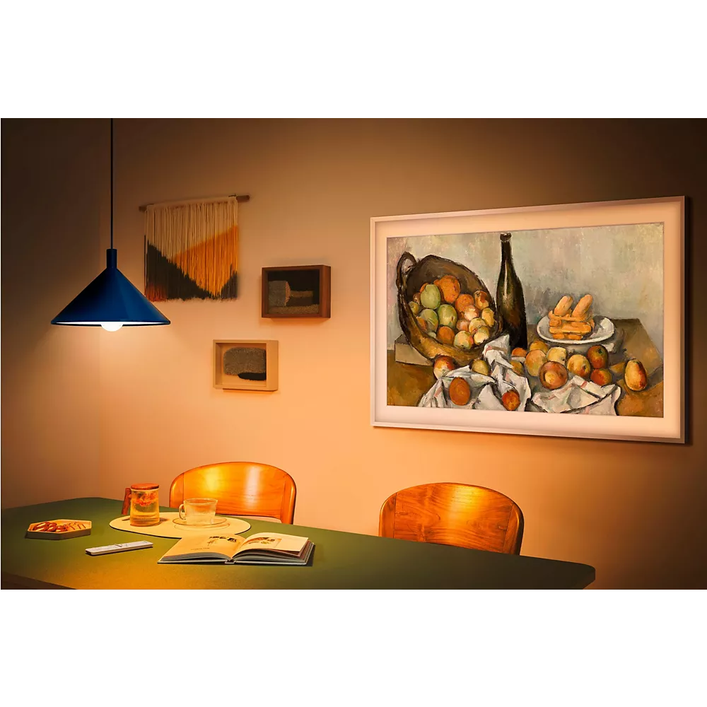 Un coin repas confortable avec un éclairage chaleureux, des œuvres d'art murales - dont l'élégant SAMSUNG TQ55LS03F THE FRAME - et une table garnie de livres, d'un verre et d'un bocal.