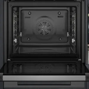 Un four SIEMENS HB472G0B3F ouvert, à l'intérieur sombre, trône à côté d'un plan de travail en marbre orné de décorations et d'un livre de cuisine.