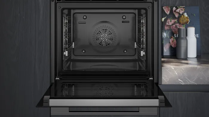 Un four SIEMENS HB472G0B3F ouvert, à l'intérieur sombre, trône à côté d'un plan de travail en marbre orné de décorations et d'un livre de cuisine.
