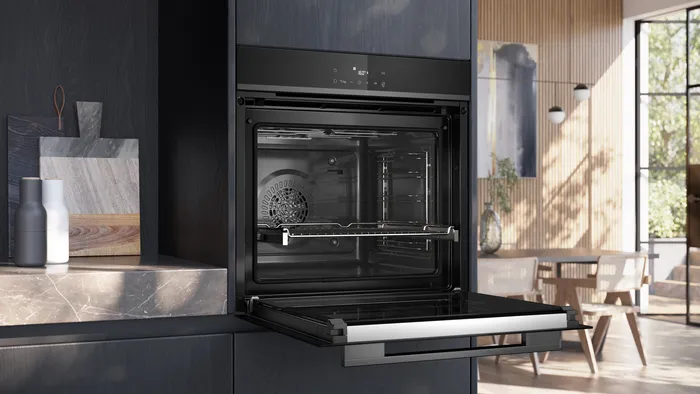 Le four encastrable SIEMENS HB472G0B3F est présenté avec sa porte ouverte dans une cuisine élégante remplie de lumière naturelle et d'élégantes touches de bois.