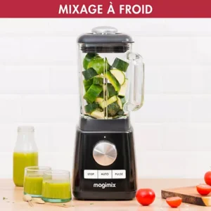 Un mixeur MAGIMIX 11628 avec des légumes hachés se trouve à côté de deux verres de jus vert et de tomates fraîches sur la table.