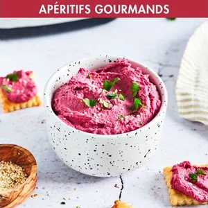 Un bol blanc de dip rose préparé avec le MAGIMIX 11630, agrémenté d'herbes, est placé à côté de crackers et de graines de sésame sur une surface claire.