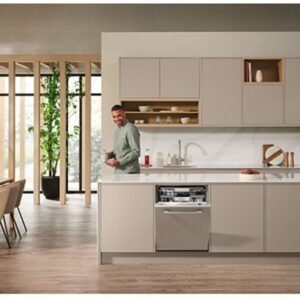 Un homme se tient dans une cuisine moderne avec des armoires beiges et un lave-vaisselle MIELE G 5863 SCVI EXCELLENCE ouvert, souriant à l'appareil photo - mettant en évidence la qualité des appareils électroménagers haut de gamme.