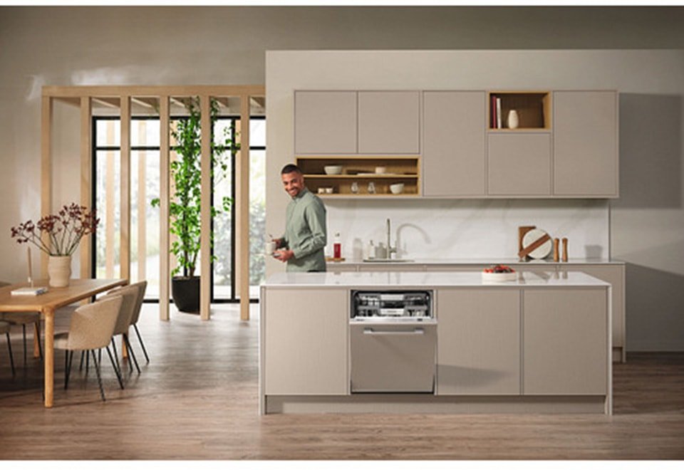 Un homme se tient dans une cuisine moderne avec des armoires beiges et un lave-vaisselle MIELE G 5863 SCVI EXCELLENCE ouvert, souriant à l'appareil photo - mettant en évidence la qualité des appareils électroménagers haut de gamme.