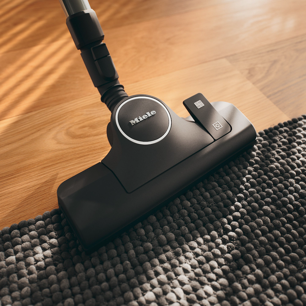 La tête d'un aspirateur Miele BOOST CX1 POWERLINE glisse sur un tapis foncé texturé sur un plancher en bois, offrant un nettoyage puissant.