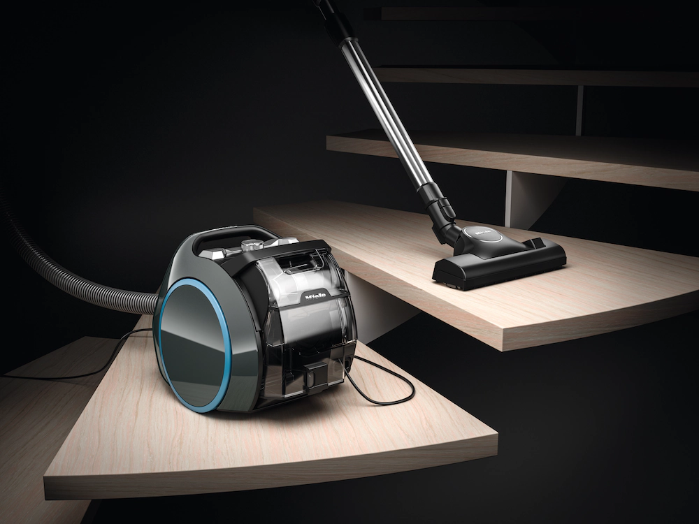 Un aspirateur Miele BOOST CX1 POWERLINE est utilisé pour nettoyer des escaliers en bois clair dans un cadre moderne et minimaliste.