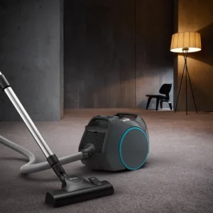 Un aspirateur Miele BOOST CX1 POWERLINE sur un sol moquetté dans une pièce moderne, avec une chaise et un lampadaire allumé en arrière-plan.