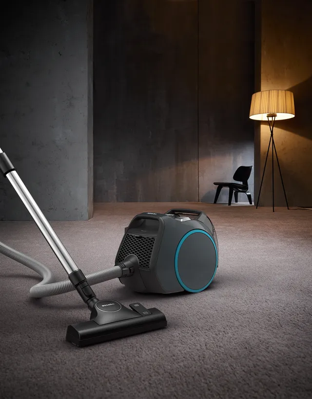 Un aspirateur Miele BOOST CX1 POWERLINE sur un sol moquetté dans une pièce moderne, avec une chaise et un lampadaire allumé en arrière-plan.
