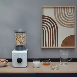 Un mixeur SMEG BLC01BLMEU contenant des noix de cajou, du lait, du miel, des amandes et de l'eau est posé sur le comptoir près d'une œuvre d'art murale abstraite. Il s'agit d'un élégant appareil de cuisine SMEG destiné à la préparation de recettes saines.