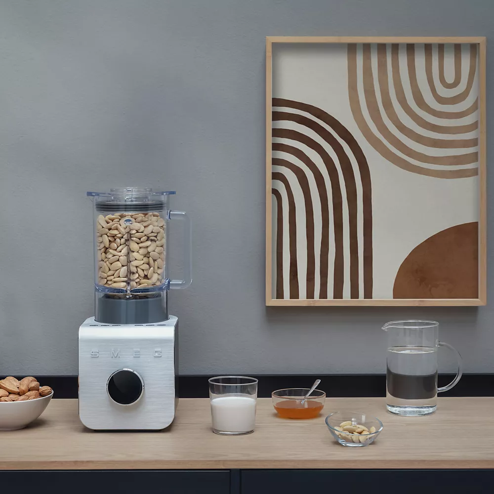Un mixeur SMEG BLC01BLMEU contenant des noix de cajou, du lait, du miel, des amandes et de l'eau est posé sur le comptoir près d'une œuvre d'art murale abstraite. Il s'agit d'un élégant appareil de cuisine SMEG destiné à la préparation de recettes saines.