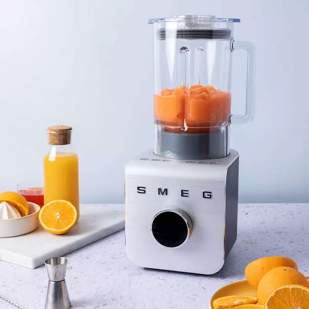 Un mixeur SMEG BLC01BLMEU est posé sur le plan de travail, rempli de smoothie à l'orange et entouré d'oranges fraîches et de jus.