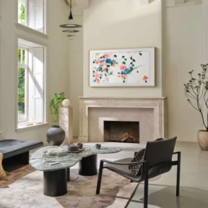 Un salon moderne avec de grandes fenêtres, de l'art abstrait au-dessus de la cheminée, un SAMSUNG TQ65LS03WF qui s'intègre parfaitement et des meubles élégants aux tons neutres.