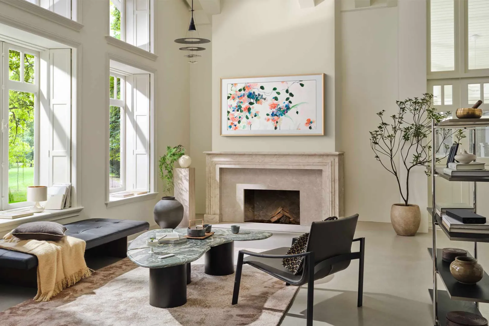 Un salon moderne avec de grandes fenêtres, de l'art abstrait au-dessus de la cheminée, un SAMSUNG TQ65LS03WF qui s'intègre parfaitement et des meubles élégants aux tons neutres.