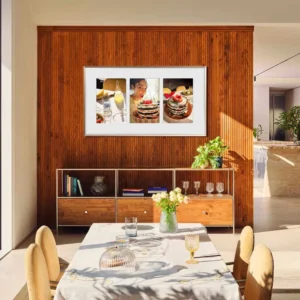 Une salle à manger moderne avec un mur en bois, une table et un tableau de crêpes au-dessus d'un buffet est rehaussée par le téléviseur SAMSUNG TQ65LS03WF, qui allie technologie et style pour un look homogène.
