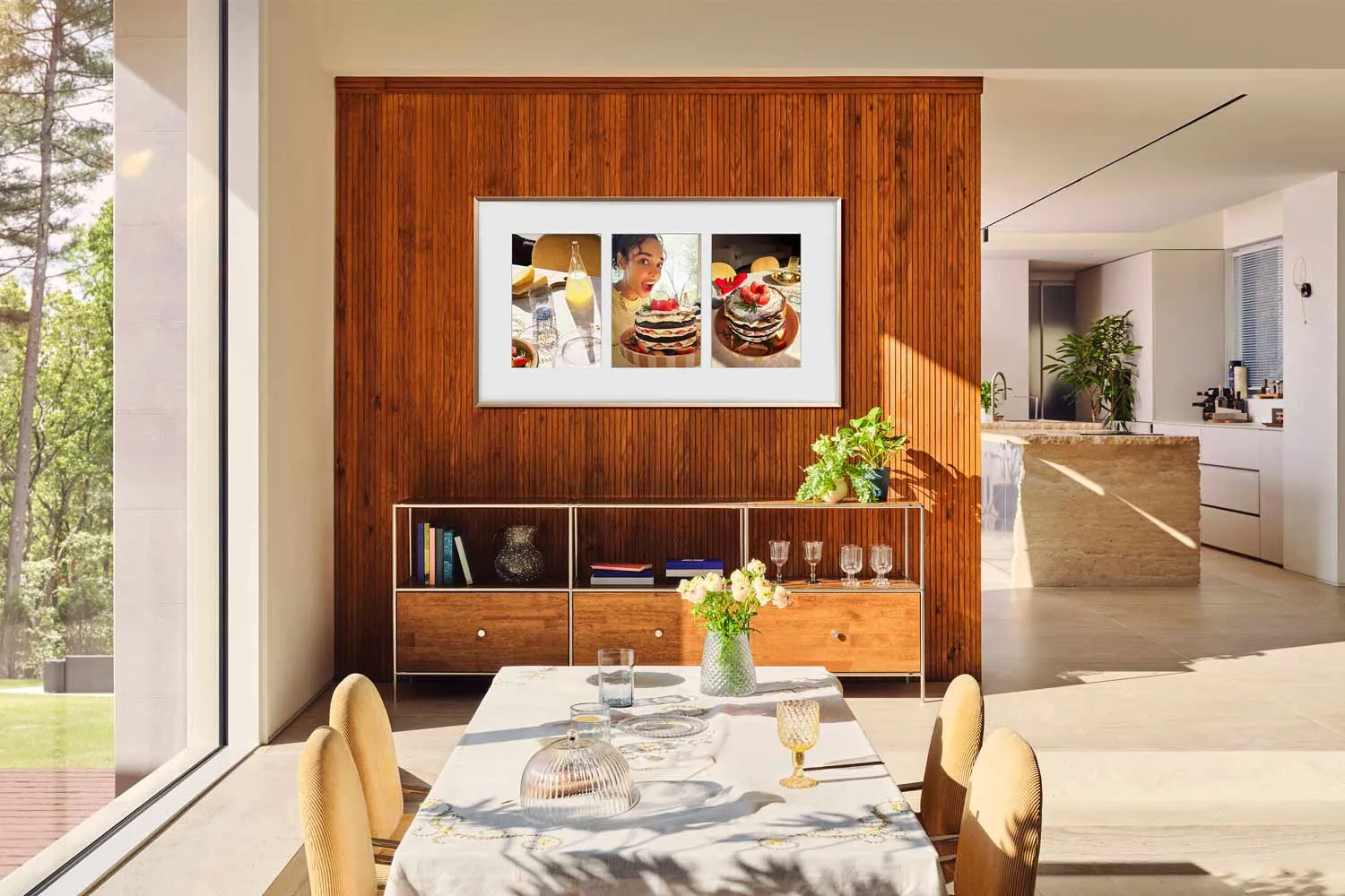 Une salle à manger moderne avec un mur en bois, une table et un tableau de crêpes au-dessus d'un buffet est rehaussée par le téléviseur SAMSUNG TQ65LS03WF, qui allie technologie et style pour un look homogène.