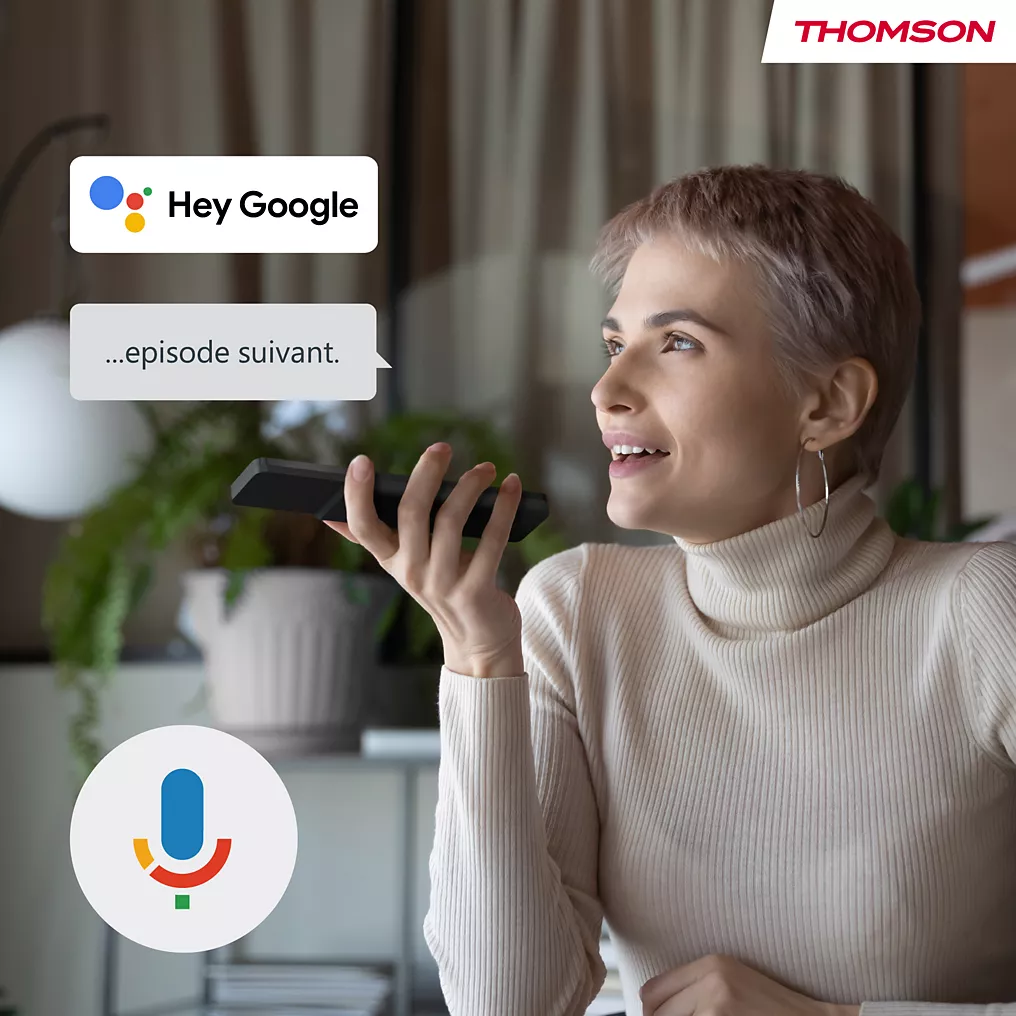 Une femme utilise la commande vocale, en disant "épisode suivant", sur son smartphone pour contrôler son THOMSON 32HA4M44, avec Google Assistant et les logos de Thomson en vue.
