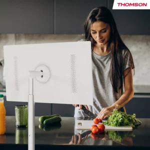 Une femme coupe des légumes dans une cuisine moderne tandis qu'un téléviseur THOMSON 32HA4M44 placé sur un support au premier plan affiche un écran blanc net.