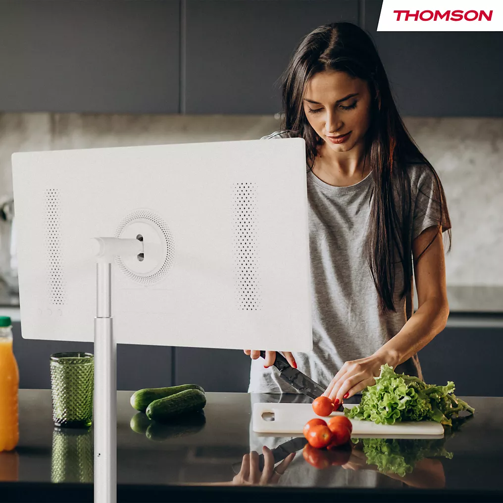 Une femme coupe des légumes dans une cuisine moderne tandis qu'un téléviseur THOMSON 32HA4M44 placé sur un support au premier plan affiche un écran blanc net.