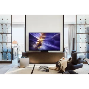 Salon moderne avec un grand téléviseur SAMSUNG TQ77s92f, un fauteuil noir, une couverture et des livres, avec une vue de la ville en arrière-plan.