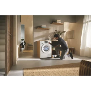 Une personne est agenouillée devant un lave-linge et un sèche-linge à pompe à chaleur MIELE TSC643WP dans une buanderie moderne et ensoleillée.