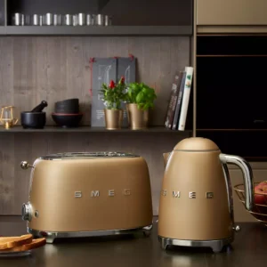La bouilloire dorée SMEG KLF03CHMEU est posée sur le plan de travail d'une cuisine, à côté du grille-pain assorti, avec des plantes et des livres de cuisine en arrière-plan.