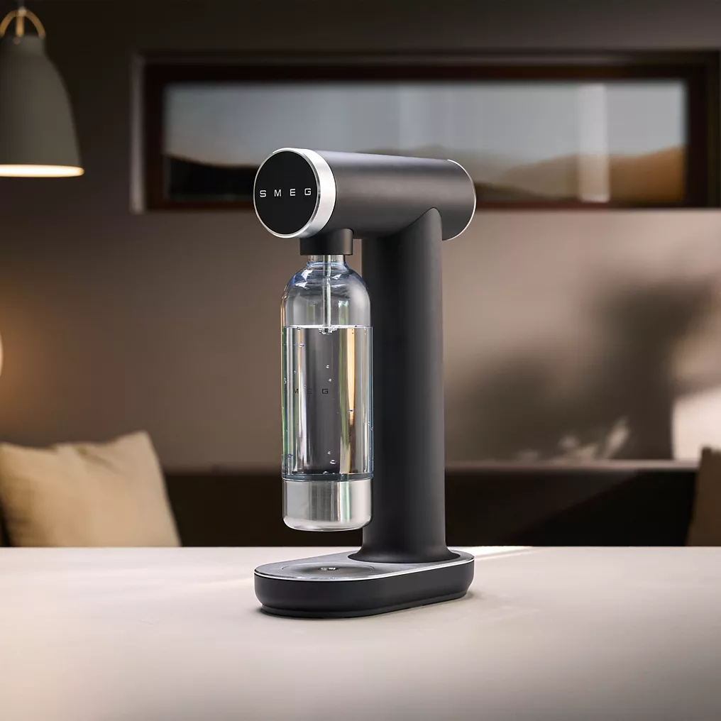 Une machine à soda SMEG SKC01BLM NOIR MAT avec une bouteille transparente est posée sur le comptoir d'une cuisine moderne.