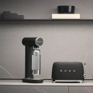 Une machine à soda SMEG SKC01BLM NOIR MAT et un grille-pain sont placés sur un plan de travail gris avec des étagères et des ustensiles de cuisine exposés au-dessus.