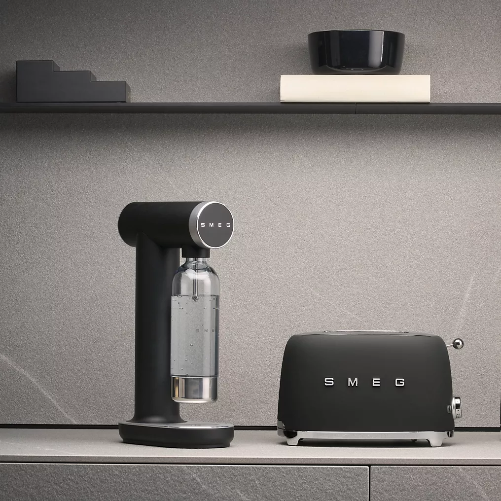 Une machine à soda SMEG SKC01BLM NOIR MAT et un grille-pain sont placés sur un plan de travail gris avec des étagères et des ustensiles de cuisine exposés au-dessus.