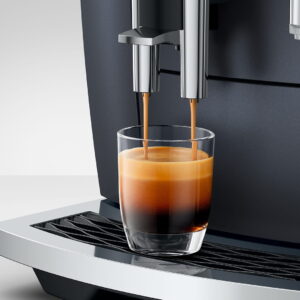 Gros plan d'une machine à café JURA E8 COSMIC BLACK (ED) versant un espresso frais dans une tasse en verre transparent sur son plateau d'égouttage en métal.