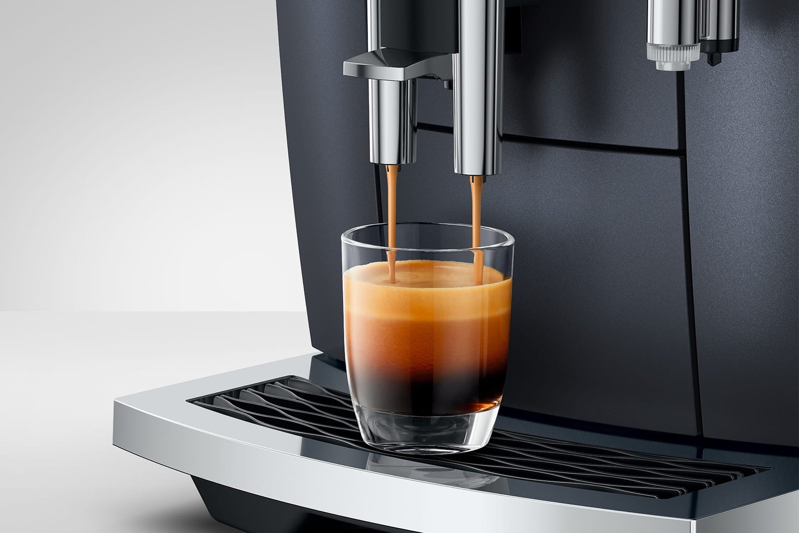 Gros plan d'une machine à café JURA E8 COSMIC BLACK (ED) versant un espresso frais dans une tasse en verre transparent sur son plateau d'égouttage en métal.