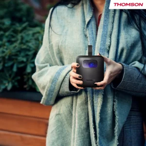 Une personne portant un châle tient un projecteur compact noir THOMSON VEGA PG35B à l'extérieur, à côté de plantes vertes.