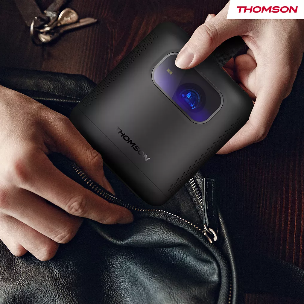 Les mains placent le projecteur compact THOMSON VEGA PG35B dans un sac noir, avec les clés visibles à proximité.