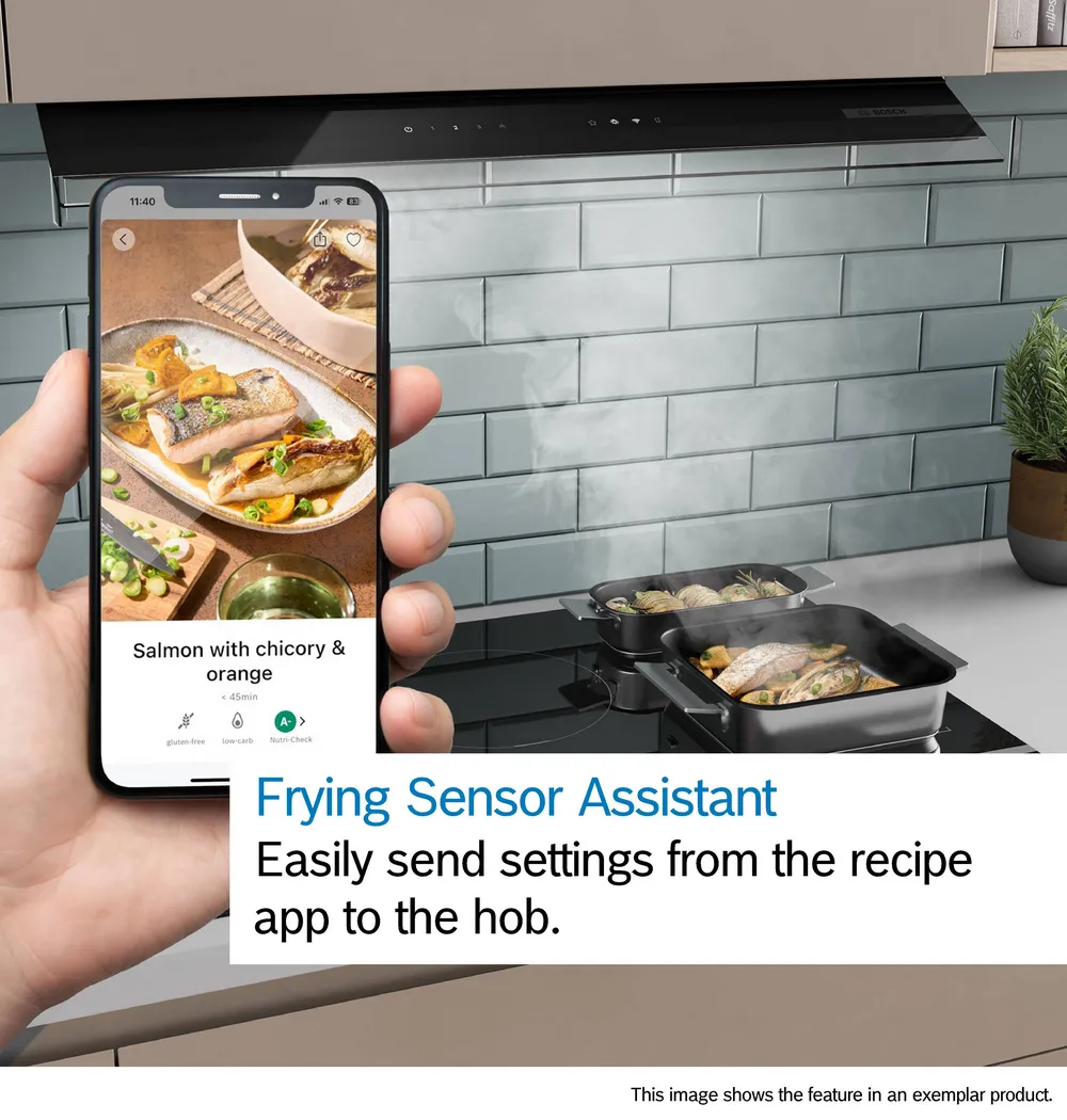 Une main tient un téléphone avec une recette de saumon devant la table de cuisson BOSCH PVW89BHC1E, où le saumon grésille dans une poêle.