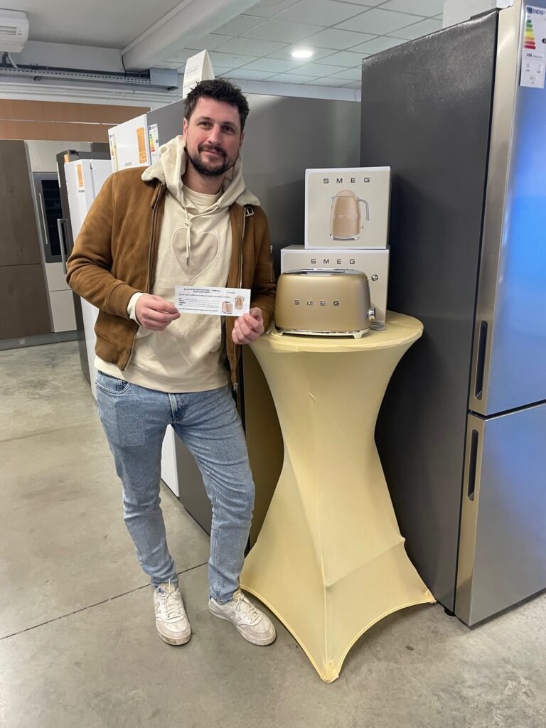 Un homme se tient à l'intérieur près d'un réfrigérateur, tenant un prospectus à côté d'un grille-pain SMEG doré et d'un plat LE CREUSET sur une table couverte.