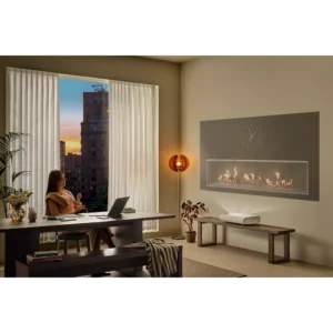 Une femme travaille sur son ordinateur portable avec un SAMSUNG SP-PLU7D à côté d'elle à un bureau dans un salon moderne, éclairé par un coucher de soleil à travers la fenêtre et la lueur d'une cheminée allumée.