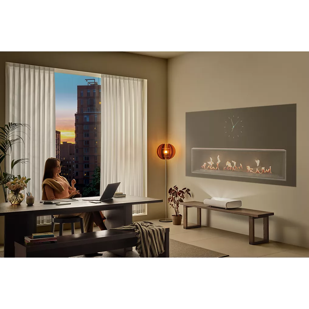 Une femme travaille sur son ordinateur portable avec un SAMSUNG SP-PLU7D à côté d'elle à un bureau dans un salon moderne, éclairé par un coucher de soleil à travers la fenêtre et la lueur d'une cheminée allumée.