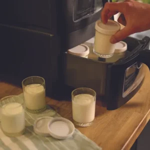 Une main place un petit pot de yaourt dans le FRIFRI AIRFRYER DUO SLIM, les pots et les couvercles étant posés sur une serviette rayée à proximité.