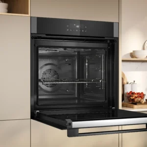 Le four encastrable NEFF B2ACJ7AG0 est présenté avec sa porte ouverte dans une cuisine moderne avec des armoires et des étagères neutres.