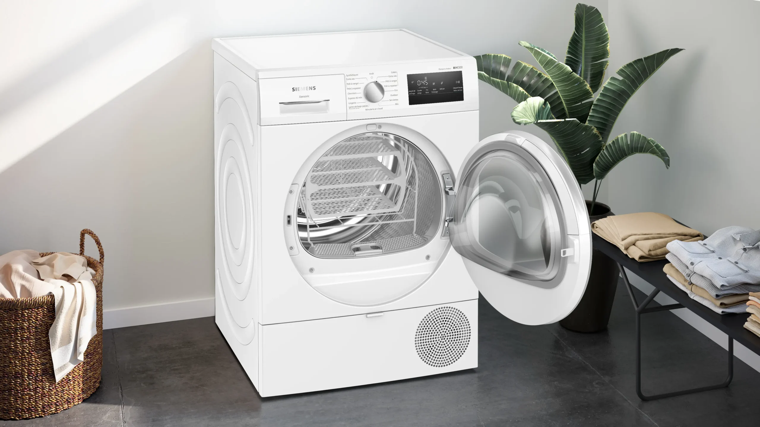 Un sèche-linge SIEMENS WT45H00MFR blanc avec sa porte ouverte est placé à côté d'une plante et de linge plié dans une pièce lumineuse.
