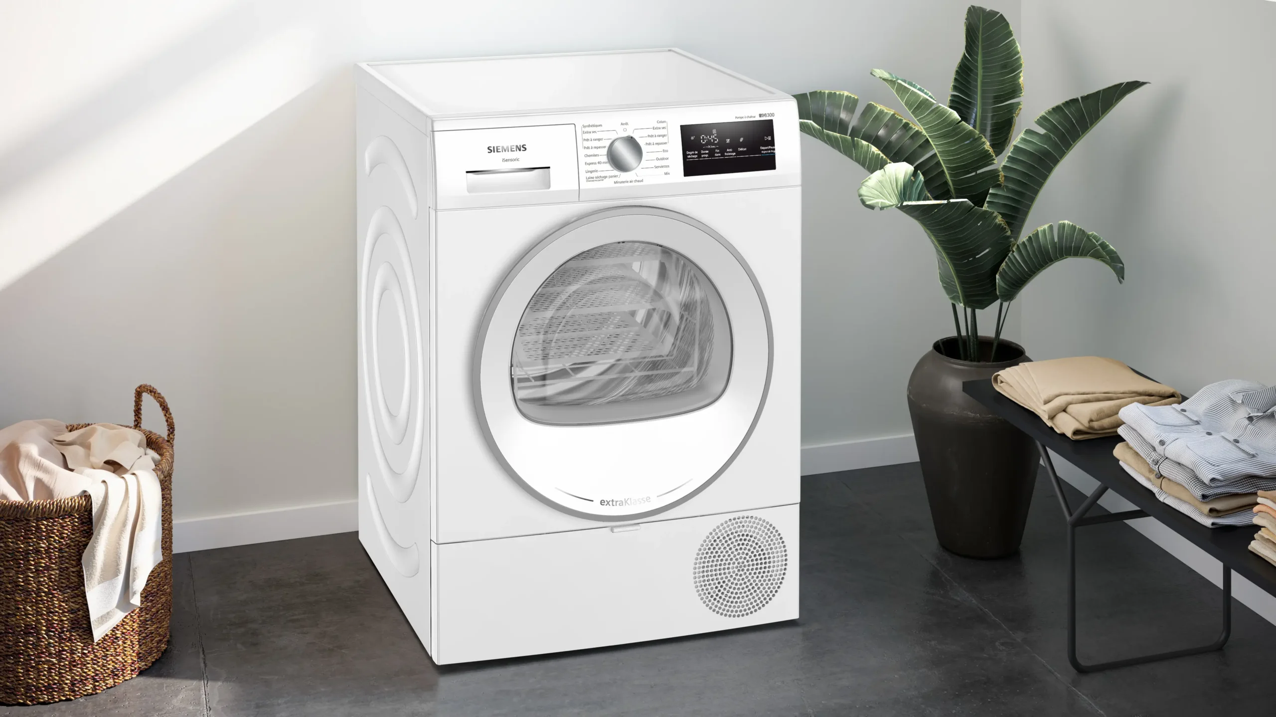Un lave-linge SIEMENS WT45H00MFR à chargement frontal de couleur blanche trône dans une buanderie moderne, entouré d'une plante, d'un panier à linge et de vêtements soigneusement pliés.