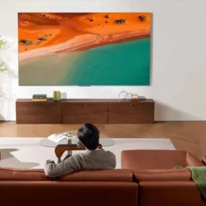 Une personne se détend sur un canapé marron, en regardant une scène de plage sur le téléviseur TCL 98C8K dans un salon moderne.