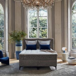 Chambre à coucher élégante avec de grandes fenêtres, un lustre, un lit gris 160x200 équipé d'un matelas EPEDA GALACTIQUE 2 160x200, une chaise bleue, un tapis rond et une vue verdoyante à l'extérieur.