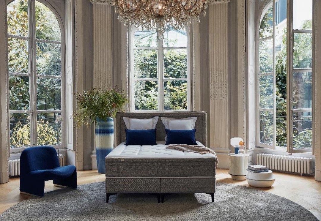 Chambre à coucher élégante avec de grandes fenêtres, un lustre, un lit gris 160x200 équipé d'un matelas EPEDA GALACTIQUE 2 160x200, une chaise bleue, un tapis rond et une vue verdoyante à l'extérieur.