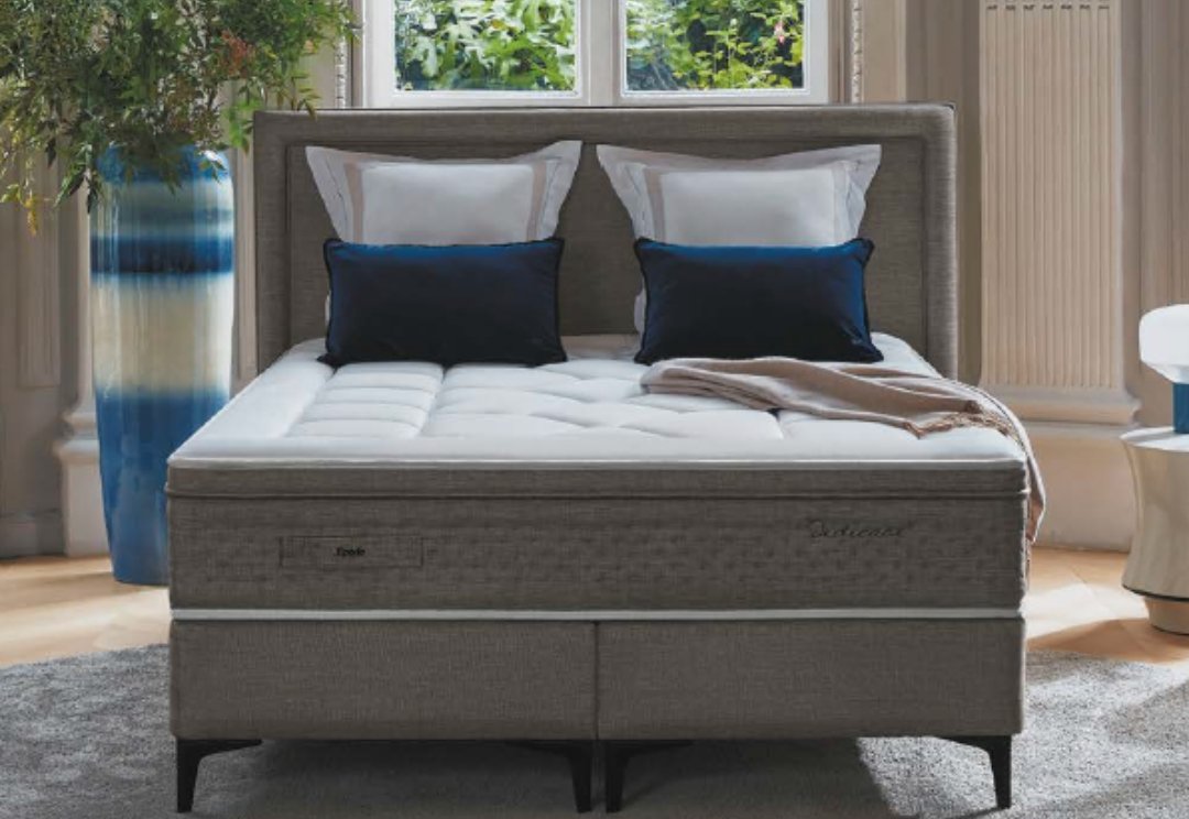 Un lit bien fait avec un matelas EPEDA GALACTIQUE 2 160x200, quatre oreillers, un jeté beige et une tête de lit tapissée grise dans une chambre lumineuse.