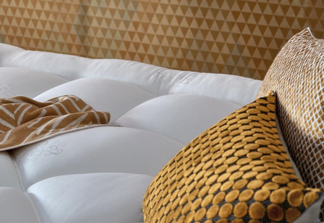 Gros plan sur le matelas EPEDA GALACTIQUE 2 160x200, dont la surface est touffetée en blanc, et qui est agrémenté d'oreillers à motifs jaunes et dorés et d'une couverture douillette.